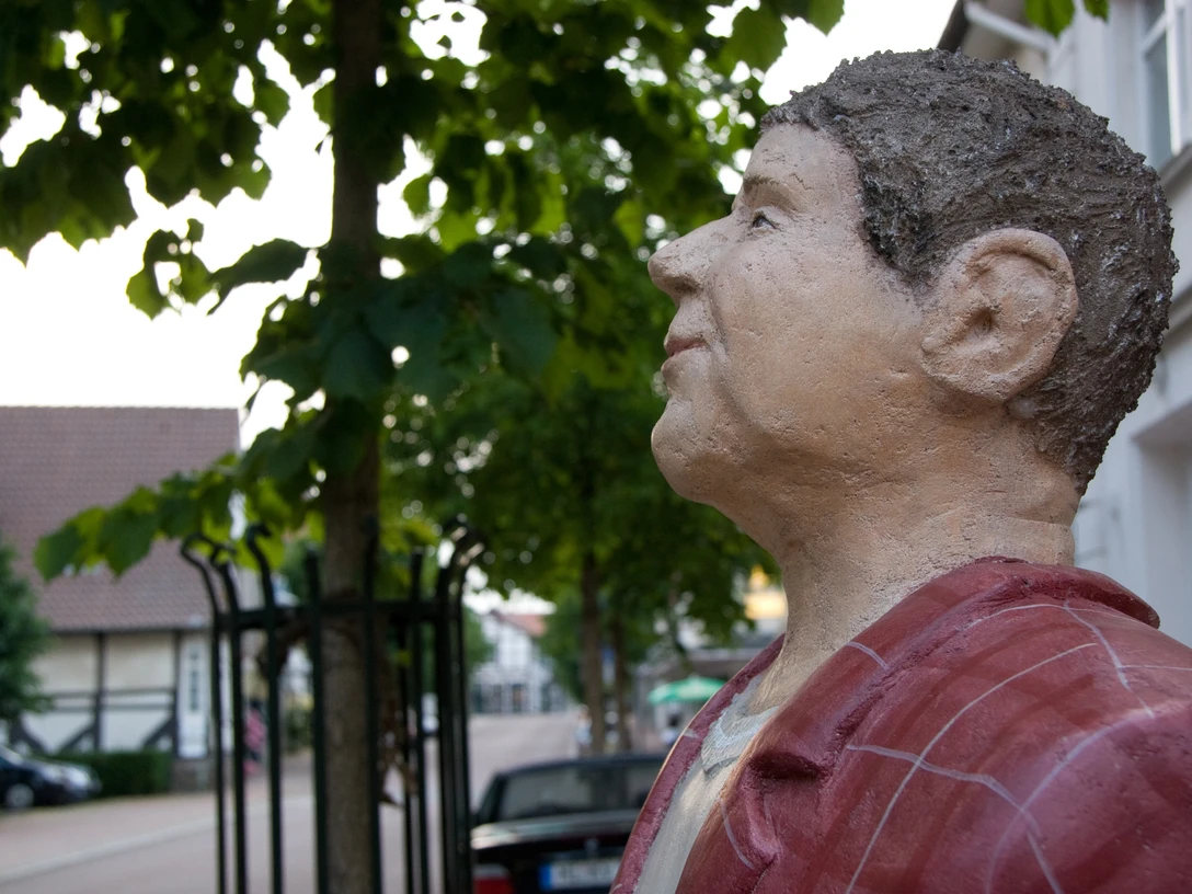 Lechnerfigur "Herr Meier" unter einem Baum, mit Fachwerkhäusern im Hintergrund.