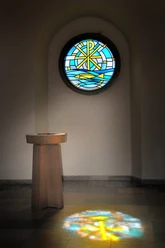 Kirche Bierde Taufstein Ein Taufstein vor einem Rundfenster, dessen buntes Glas ein farbenfrohes Muster auf den Boden projiziert.