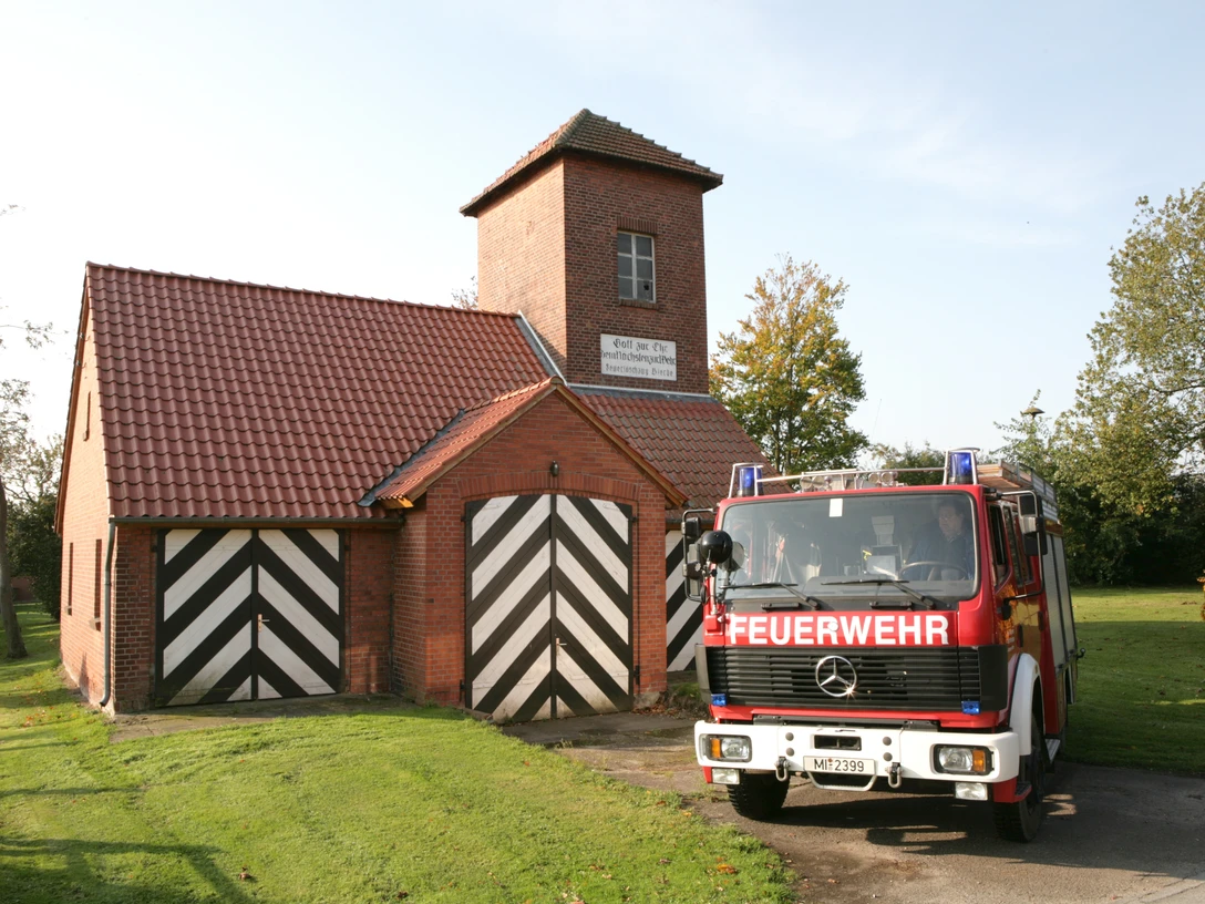 Feuerwehrgerätehaus Bierde Ein rotes Feuerwehrfahrzeug vor einem Backsteingebäude mit typischen schwarz-weißen Toren.