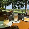 Gastronomie an der Weser in Buchholz Käsekuchen und Latte Macchiato auf Holztisch, umgeben von Bäumen mit Blick auf grüne Wiesen.