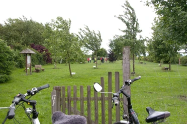 Zwei Fahrräder stehen vor einem Holztor, dahinter eine grüne Wiese mit jungen Bäumen und Spaziergängern.