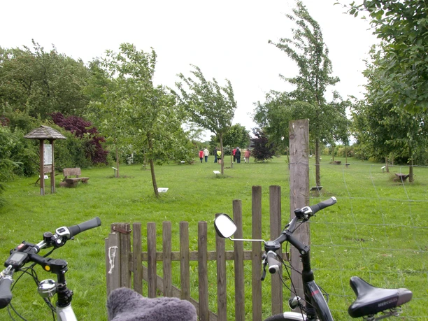 Zwei Fahrräder stehen vor einem Holztor, dahinter eine grüne Wiese mit jungen Bäumen und Spaziergängern.