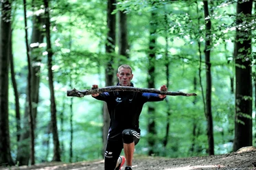 Fitness-Wanderung mit Trainer Robert Dreisiebner Trainer Robert Dreisiebner führt eine Übung im Wald aus, umgeben von hohen Bäumen und grünem Laub.