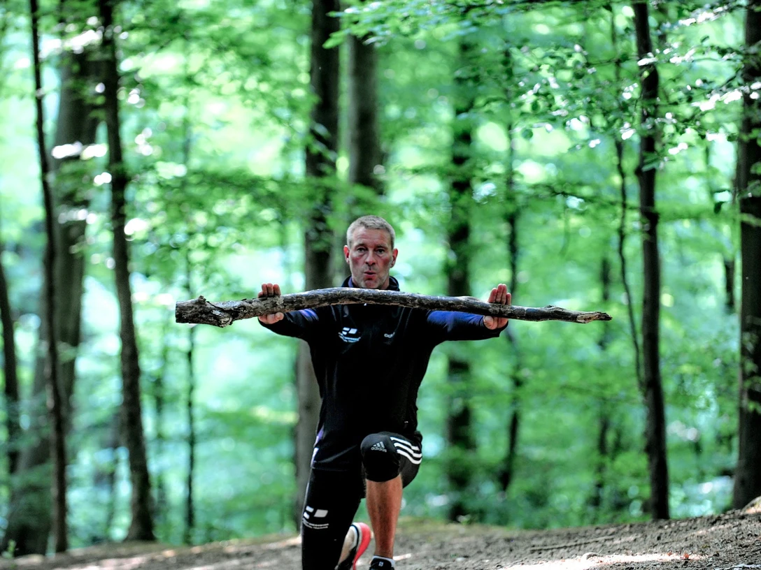 Fitness-Wanderung mit Trainer Robert Dreisiebner Trainer Robert Dreisiebner führt eine Übung im Wald aus, umgeben von hohen Bäumen und grünem Laub.