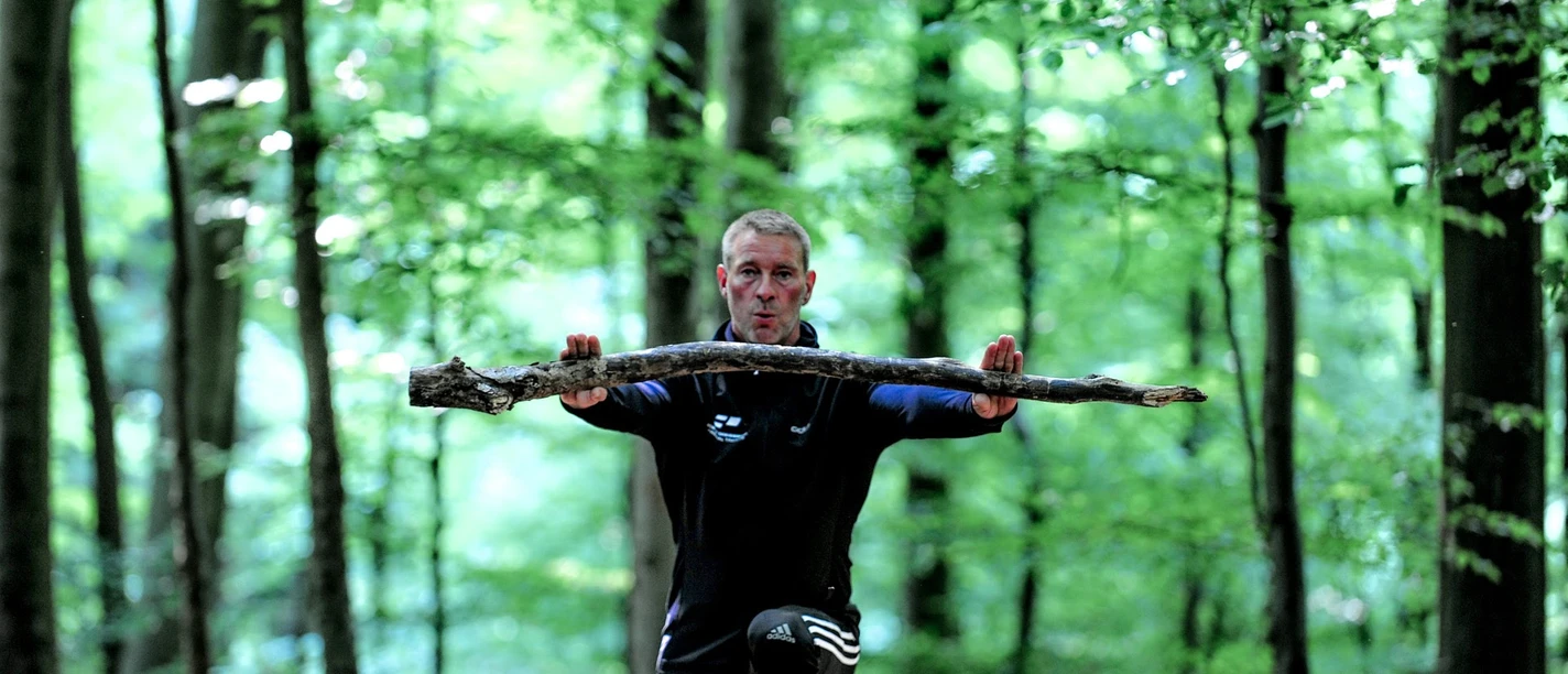 Fitness-Wanderung mit Trainer Robert Dreisiebner Trainer Robert Dreisiebner führt eine Übung im Wald aus, umgeben von hohen Bäumen und grünem Laub.