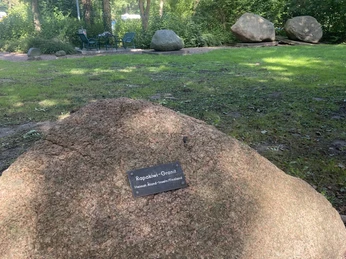 Ein großer Felsbrocken mit einer kleinen Gedenktafel darauf steht in einem Waldgebiet mit grünen Bäumen.