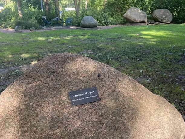 Ein großer Felsbrocken mit einer kleinen Gedenktafel darauf steht in einem Waldgebiet mit grünen Bäumen.