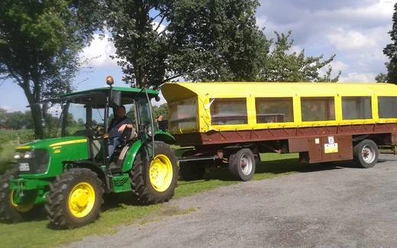 Wiehes_Planwagen_1_c33e30a3c9.jpg Grüner Traktor zieht einen gelben Planwagen über einen gepflasterten Weg, Bäume im Hintergrund.