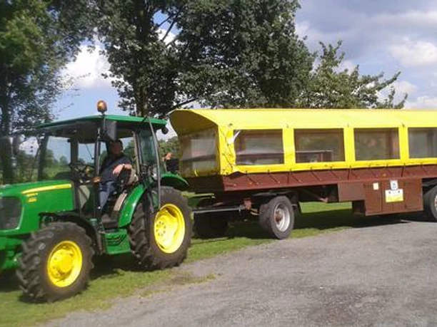 Wiehes_Planwagen_1_c33e30a3c9.jpg Grüner Traktor zieht einen gelben Planwagen über einen gepflasterten Weg, Bäume im Hintergrund.