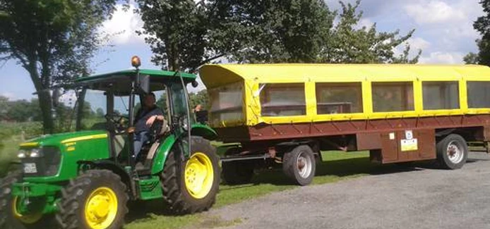 Wiehes_Planwagen_1_c33e30a3c9.jpg Grüner Traktor zieht einen gelben Planwagen über einen gepflasterten Weg, Bäume im Hintergrund.