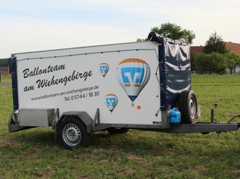 Anhänger mit Werbung des Ballonteams am Wiehengebirge auf einer grünen Wiese, umgeben von Bäumen.