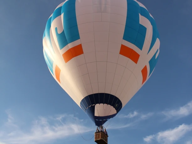 Heißluftballon schwebt friedlich über einer hügeligen Landschaft, vor einem blauen Himmel mit Wolken.