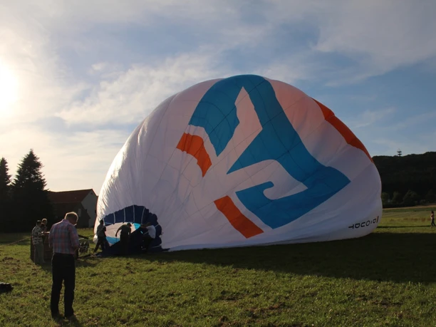 Heißluftballon am Boden mit Menschen beim Vorbereiten, im Hintergrund Hügel und Häuser unter blauem Himmel.
