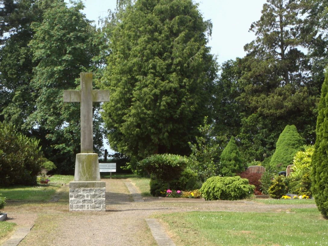 Ein großzügiger Friedhof mit einem steinernen Kreuz, umgeben von grünen Bäumen und gepflegten Pflanzen.