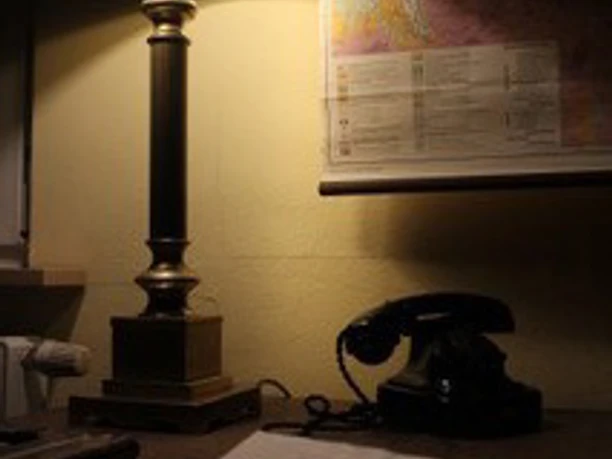 EscapeRoom.jpg Ein Schreibtisch mit alter Telefonapparatur, Notizblock, Tischlampe und Landkarte an der Wand.