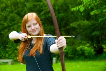 Bogenschießen am Neanderthal Museum in Mettmann Person mit rotem Haar spannt einen Bogen im Freien vor grünem Hintergrund.
