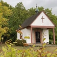 Kleine Kapelle mit Kreuz im Grünen, umgeben von farbenfrohem Herbstlaub und bewölktem Himmel.