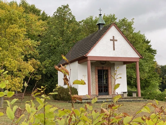 Kleine Kapelle mit Kreuz im Grünen, umgeben von farbenfrohem Herbstlaub und bewölktem Himmel.