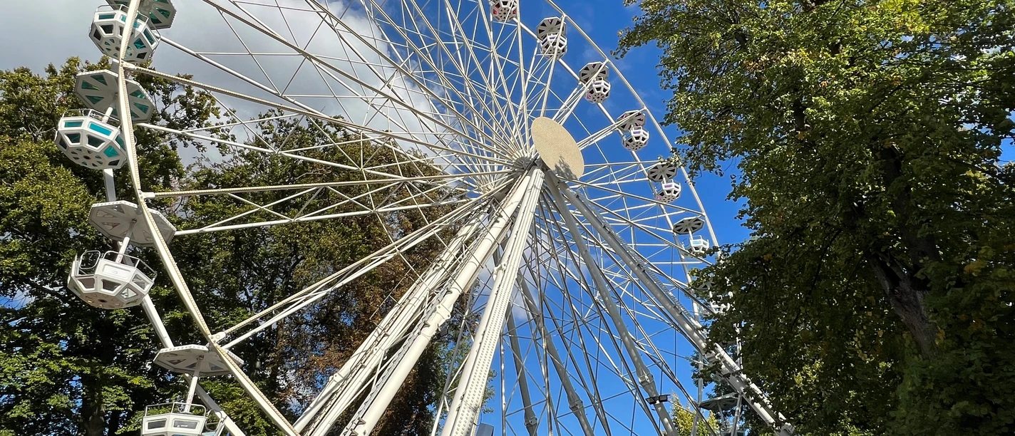 Riesenrad A white Ferris wheel rises against a clear blue sky, surrounded by green trees.