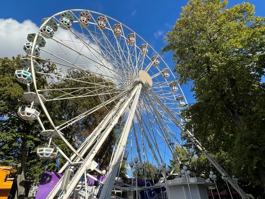 Riesenrad Ein weißes Riesenrad erhebt sich vor einem klaren blauen Himmel, umgeben von grünen Bäumen.A white Ferris wheel rises against a clear blue sky, surrounded by green trees.Et hvidt pariserhjul rejser sig mod en klar blå himmel, omgivet af grønne træer.Een wit reuzenrad rijst op tegen een helderblauwe hemel, omringd door groene bomen.