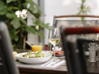 Auf der Terrasse im Hotel und Restaurant Haus Fabry in Hilden Auf einem Tisch stehen ein Salatteller, ein Glas Weißwein und ein Glas Orangensaft, umgeben von Grünpflanzen.