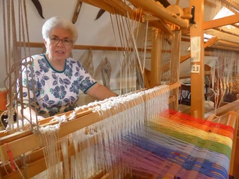 Webstube Ilse Eine ältere Frau sitzt am Webstuhl und arbeitet an einem bunten Stoff, im Raum hängen weitere Webarbeiten.
