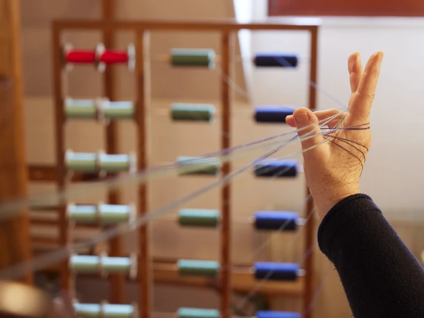 Ein Handwebstuhl mit Garnrollen, fokussiert auf eine Hand, die bunte Fäden führt.