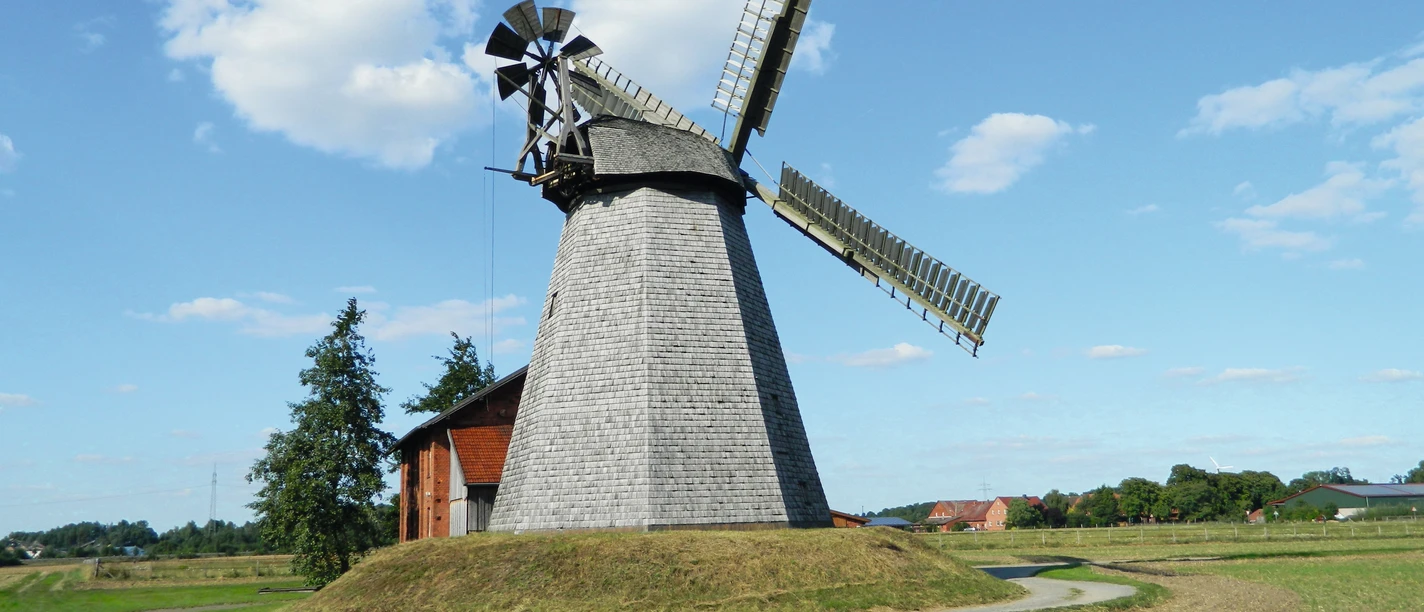 Windmühle Bierde mit grauem Holzschindeldach und Flügeln in einer weiten, grünen Landschaft.