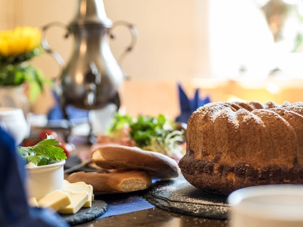 Bergische Kaffeetafel Ein gedeckter Tisch mit Gugelhupf, Brot, Butter und einer Kanne im Hintergrund.