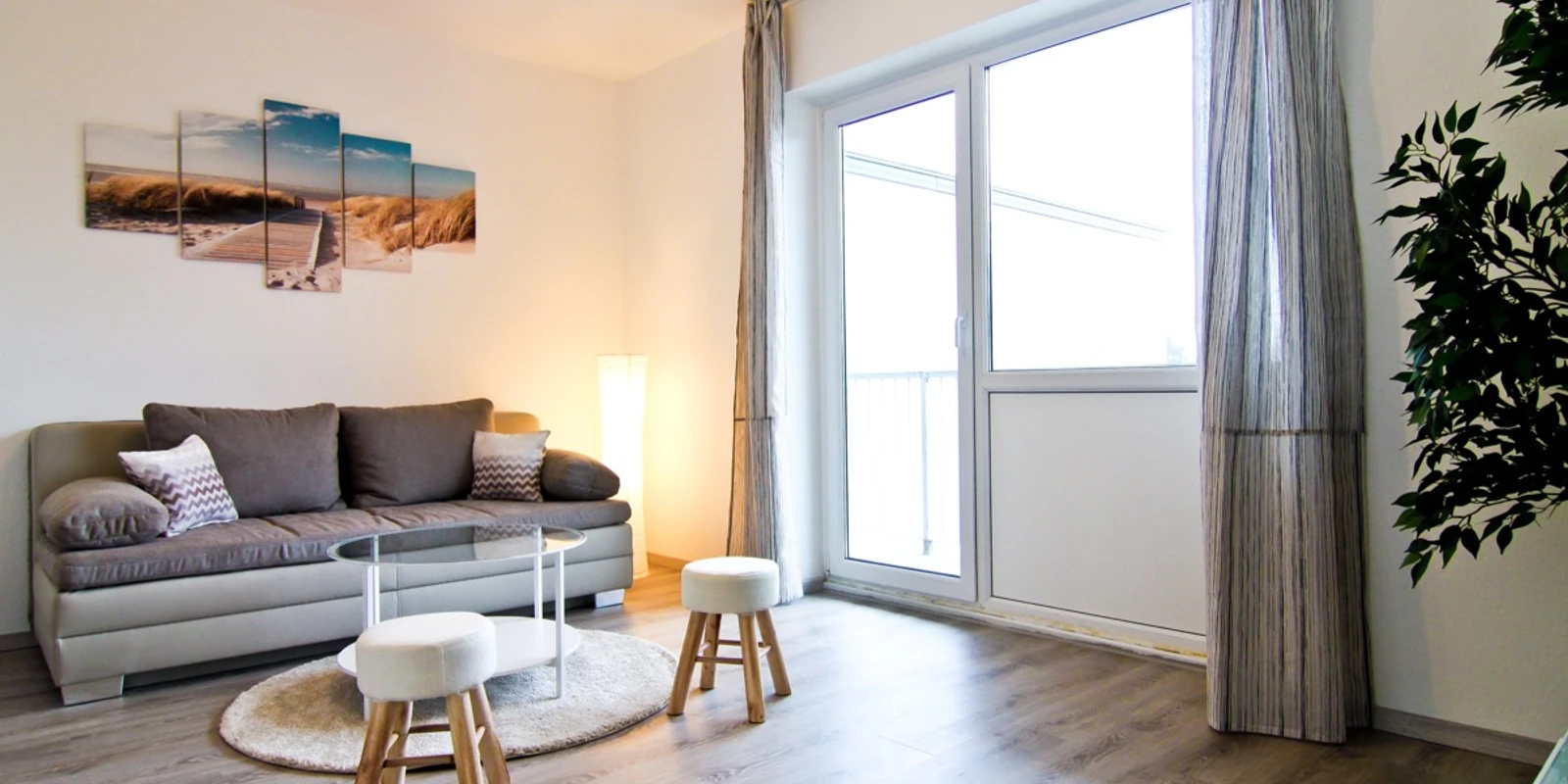 apartmondo Ferienwohnung in Heiligenhaus Helles Wohnzimmer in Ferienwohnung, ausgestattet mit grauem Sofa, Couchtisch, zwei Hockern und Wandbild.