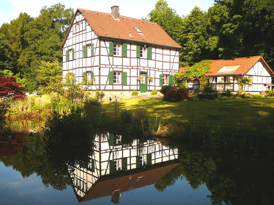Gästehaus Wahnenmühle in Erkrath Weißes Fachwerkhaus mit grünen Fensterläden am Wasser, umgeben von üppiger Vegetation in Erkrath.