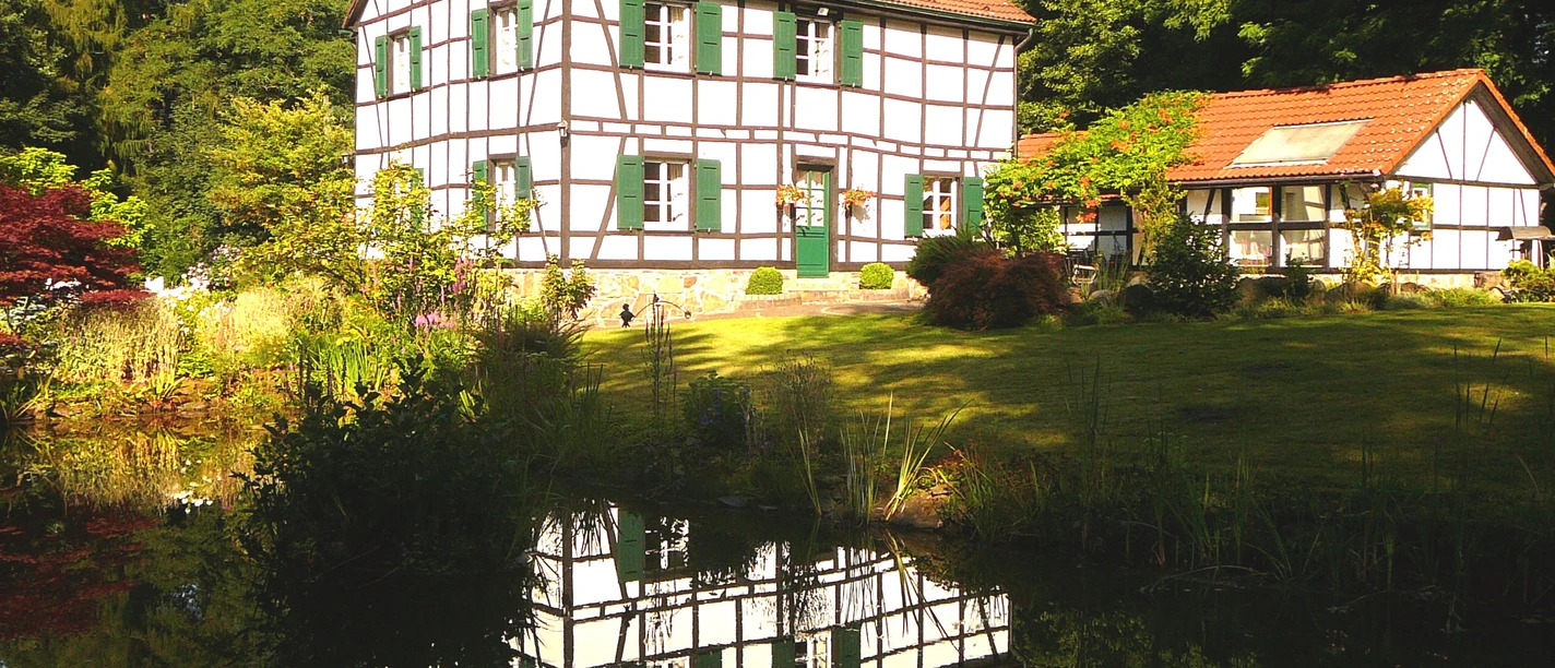 Gästehaus Wahnenmühle in Erkrath Weißes Fachwerkhaus mit grünen Fensterläden am Wasser, umgeben von üppiger Vegetation in Erkrath.