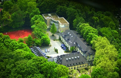 Waldhotel Heiligenhaus Luftaufnahme eines von Wald umgebenen Hotels mit Tennisplatz und moderner Gebäudeerweiterung.