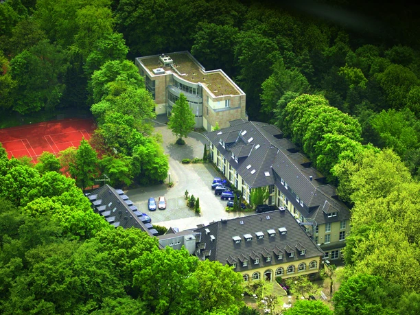 Waldhotel Heiligenhaus Luftaufnahme eines von Wald umgebenen Hotels mit Tennisplatz und moderner Gebäudeerweiterung.