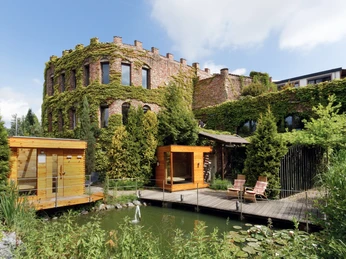 Land Gut Höhne in Mettmann Ein mit Efeu bewachsenes Gebäude neben einem Teich, umgeben von saftigem, grünem Garten.