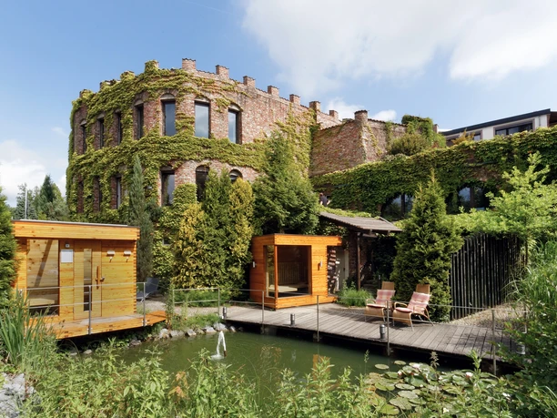 Land Gut Höhne in Mettmann Ein mit Efeu bewachsenes Gebäude neben einem Teich, umgeben von saftigem, grünem Garten.