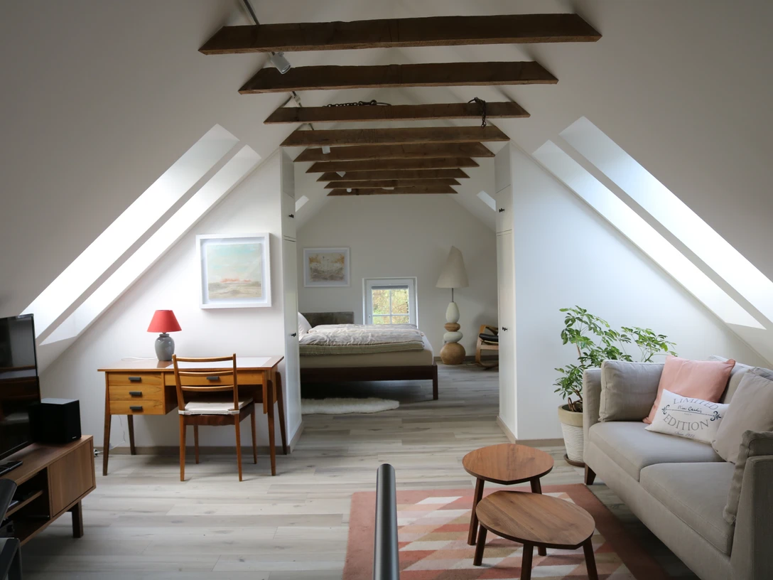 Gut Steineshof in Mettmann Helles Dachgeschoss mit sichtbaren Balken, Schreibtisch, Sofa und Bett im Hintergrund, moderner Stil.