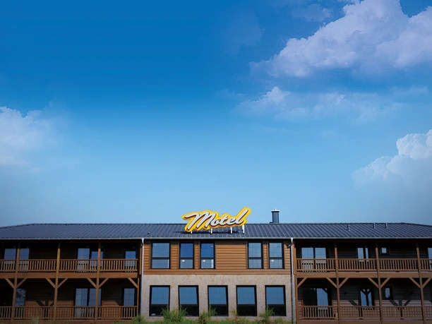 Road Stop in Mettmann Zweistöckiges Motel mit Holzbalkonen, blauer Himmel und dezentes "Motel"-Schild im Vordergrund.