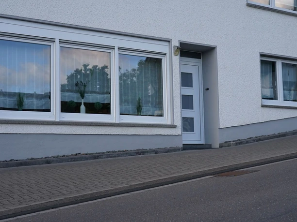 Außenansicht Hausfront mit zwei Fenstern und Eingangstür, an einer leicht geneigten Straße gelegen.