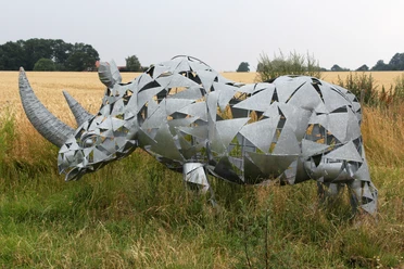 Metallic rhinoceros sculpture with geometric shapes stands in a meadow in front of a field of grain.