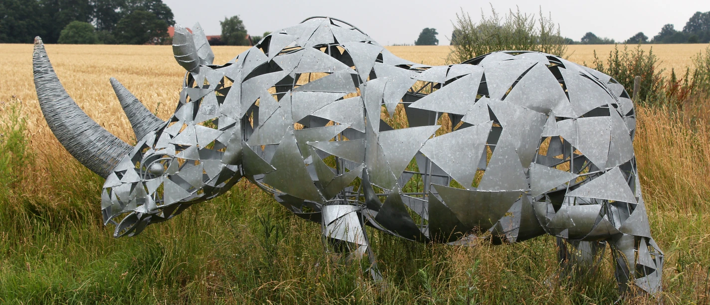 Skulptur Nashorn Metallic rhinoceros sculpture with geometric shapes stands in a meadow in front of a field of grain.