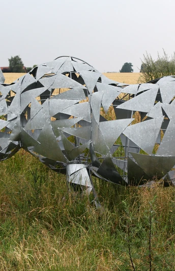 Skulptur Nashorn Metallische Nashornskulptur mit geometrischen Formen steht auf einer Wiese vor einem Getreidefeld.