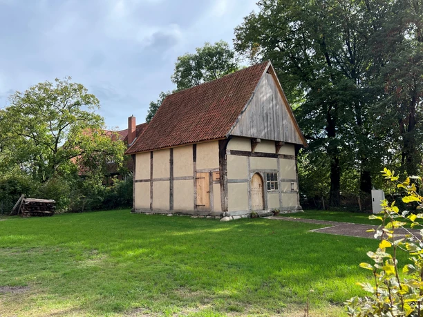 A half-timbered house with a wooden roof, surrounded by green meadows and trees in the sunlight.