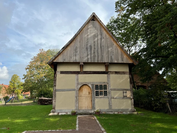 Ein altes Fachwerkgebäude mit Satteldach und Holztür, umgeben von Bäumen und einem gepflasterten Weg.