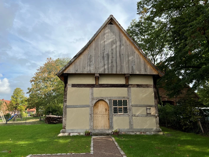 Pastorenhaus Martfeld Ein altes Fachwerkgebäude mit Satteldach und Holztür, umgeben von Bäumen und einem gepflasterten Weg.