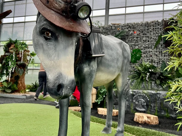 Grubenesel „Pablo“ Grauer Esel mit Bergarbeiterhelm und Lampe in einer grünen, botanisch gestalteten Umgebung.
