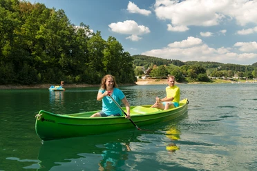 Freizeitcamp Aggertalsperre Kanufahrer auf einem grünen See umgeben von bewaldeten Hügeln unter einem blauem Himmel mit weißen Wolken.
