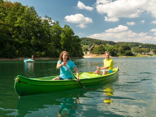 Freizeitcamp Aggertalsperre Kanufahrer auf einem grünen See umgeben von bewaldeten Hügeln unter einem blauem Himmel mit weißen Wolken.