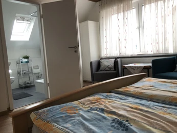 Doppelzimmer Ein Doppelzimmer mit hellem Holzinterieur, Fenster mit Vorhängen, kleinem Sofa und offenem Badzugang.