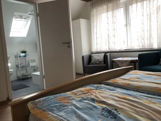 Doppelzimmer Ein Doppelzimmer mit hellem Holzinterieur, Fenster mit Vorhängen, kleinem Sofa und offenem Badzugang.